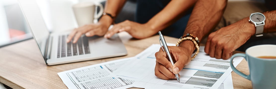 Hands working on a laptop and filling out financial documents spread across a desk with coffee nearby.